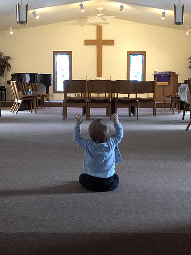 a toddler sits in the back of the sanctuary with his hands raised